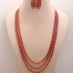 Pode incluir: Um conjunto de colar e brincos dourados. O colar apresenta vários fios de pequenas pedras redondas de cor rubi. Os brincos a condizer são uma cascata das mesmas pedras de rubi. As joias são exibidas num manequim.