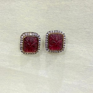 May include: A pair of square-shaped earrings with a carved pink stone set in a silver frame with a row of clear stones around the edge.