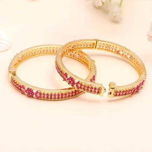 May include: Two gold-toned hinged bangles featuring a repeating floral pattern set with small, vibrant pink stones. The bracelets have a metallic finish and a secure clasp mechanism, showcasing a detailed design with rows of pink stones.