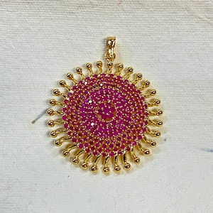 May include: A gold pendant with a circular design featuring a cluster of pink gemstones. The pendant has a sunburst-like pattern with gold accents.