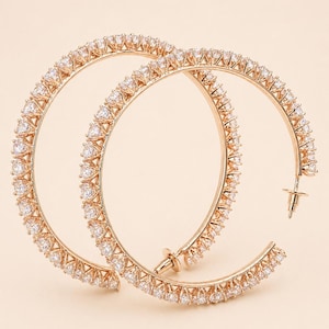 May include: A pair of rose gold hoop earrings. Each earring is encrusted with small, clear gemstones. The earrings have a classic hoop design with a post closure. The gemstones are set closely together, creating a sparkling effect.