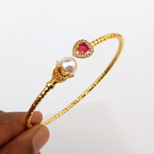 Può includere: Un braccialetto dorato con una gemma rossa a forma di cuore e una perla. Il braccialetto ha un design strutturato ed è tenuto da una mano su uno sfondo bianco. La gemma a forma di cuore è circondata da piccole pietre trasparenti.