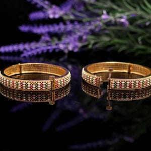 May include: Two gold bangles with intricate detailing. Each bangle features a pattern of white, red, and green accents. The bangles are displayed on a reflective black surface, with a blurred purple floral background.