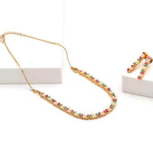 May include: A gold-toned necklace and matching pair of earrings featuring a repeating pattern of small white pearls, marquise-cut red stones, and marquise-cut green stones set in a delicate metal chain design with a lobster claw clasp.