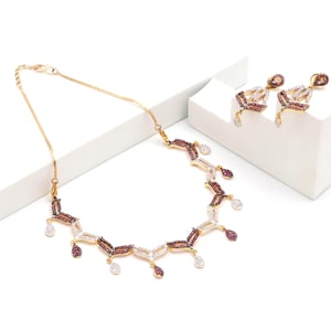 Puede incluir: Un collar de tono dorado y un par de pendientes a juego con un diseño geométrico. La joyería está engastada con pequeñas piedras blancas transparentes y piedras de color rojo oscuro dispuestas en patrones alternos, con detalles colgantes.