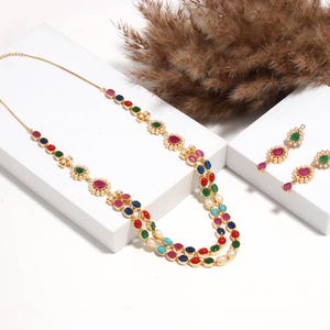 Puede incluir: Un conjunto de collar y pendientes dorados. El collar presenta dos hilos de piedras preciosas de colores, incluyendo rojo, verde, azul y blanco, engastadas en un patrón floral. Los pendientes tienen un diseño similar.