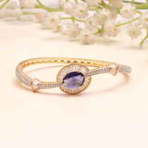 Può includere: Un bracciale rigido incernierato color oro con una gemma centrale viola dal taglio ovale, circondata da un alone di pietre chiare taglio baguette. Il bracciale è impreziosito da file di piccole pietre incastonate a pavé e dettagli.