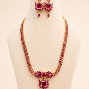 May include: A gold-toned necklace and earring set with ruby-colored gemstones. The necklace features a central pendant with a large square stone and a teardrop dangle. The earrings mirror the pendant design. The set is displayed on a mannequin.
