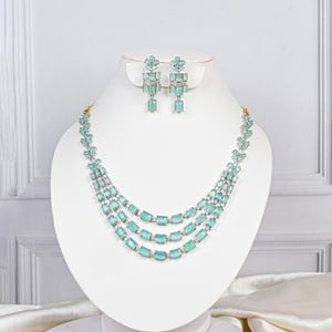 Op de afbeelding: Een sieradenset bestaande uit een ketting met drie strengen en bijpassende oorbellen. De sieraden hebben rechthoekige, lichtblauwe edelstenen gezet in een zilverkleurig metaal. De ketting wordt getoond op een witte buste.