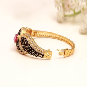 May include: A gold-tone hinged bangle bracelet featuring a black enamel section with intricate floral patterns, accented by clear pavé-set crystals and a single rectangular pink stone at the centre. The bracelet includes a secure clasp mechanism.