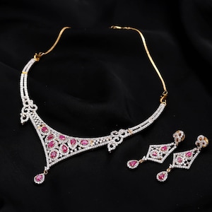 May include: A gold-toned necklace and matching pair of drop earrings featuring a central triangular pendant design. The jewelry set is adorned with numerous small clear white stones and multiple pear-shaped deep pink stones set in metal.