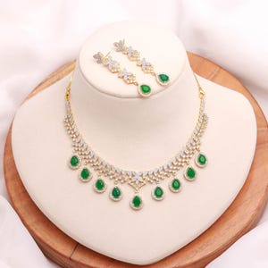 Peut inclure: Un ensemble collier et boucles d'oreilles doré avec des pierres précieuses claires et vertes. Le collier présente une série de pierres vertes en forme de goutte et de pierres claires. Les boucles d'oreilles ont un motif floral avec une seule pierre verte en forme de goutte.