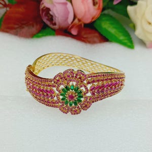 Può includere: Un bracciale rigido incernierato color oro con un motivo floreale centrale incastonato con pietre verdi taglio marquise e una pietra rossa rotonda. Il cinturino è tempestato di numerose piccole pietre rosse a motivo marquise e ha una trama.