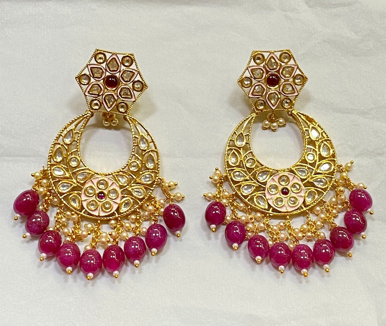 May include: A pair of gold earrings with a pink and white floral design, surrounded by small white pearls and dangling pink beads.