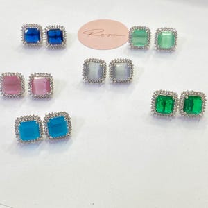 May include: A selection of square stud earrings in shades of blue, green, pink, and teal. Each pair features a central gemstone encircled by a halo of small, sparkling stones. The earrings are presented on a white surface.