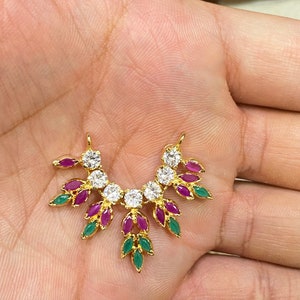 May include: A gold-toned pendant featuring a curved arrangement of clear round stones, marquise-cut pink stones, and marquise-cut green stones. The design includes two small loops at the top for attachment, with a symmetrical leaf-like pattern.