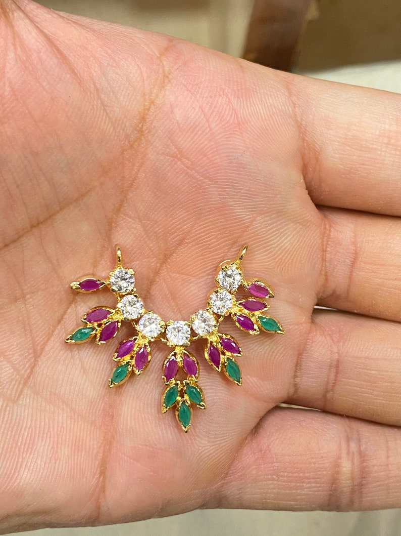 May include: A gold-toned pendant featuring a curved arrangement of clear round stones, marquise-cut pink stones, and marquise-cut green stones. The design includes two small loops at the top for attachment, with a symmetrical leaf-like pattern.