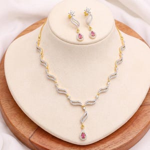 Puede incluir: Un conjunto de collar y pendientes en tono dorado con una serie de eslabones entrelazados en forma de onda incrustados con pequeñas piedras blancas. El collar incluye un colgante central, y tanto los pendientes como el colgante presentan una piedra rosa en forma de lágrima.