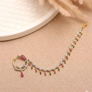 May include: Gold-toned nose ring with a floral design, featuring clear and colored gemstones. The ring has a delicate chain with alternating red and green gems. A teardrop-shaped pink gem hangs from the ring's center.