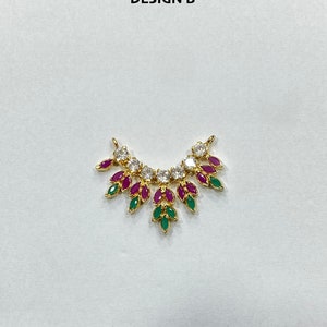 May include: A gold-toned pendant labeled DESIGN B, featuring a curved arrangement of clear round stones and marquise-cut fuchsia and green stones. The piece has two small loops at the top ends for attachment, creating a fan-like, symmetrical jewelry component.