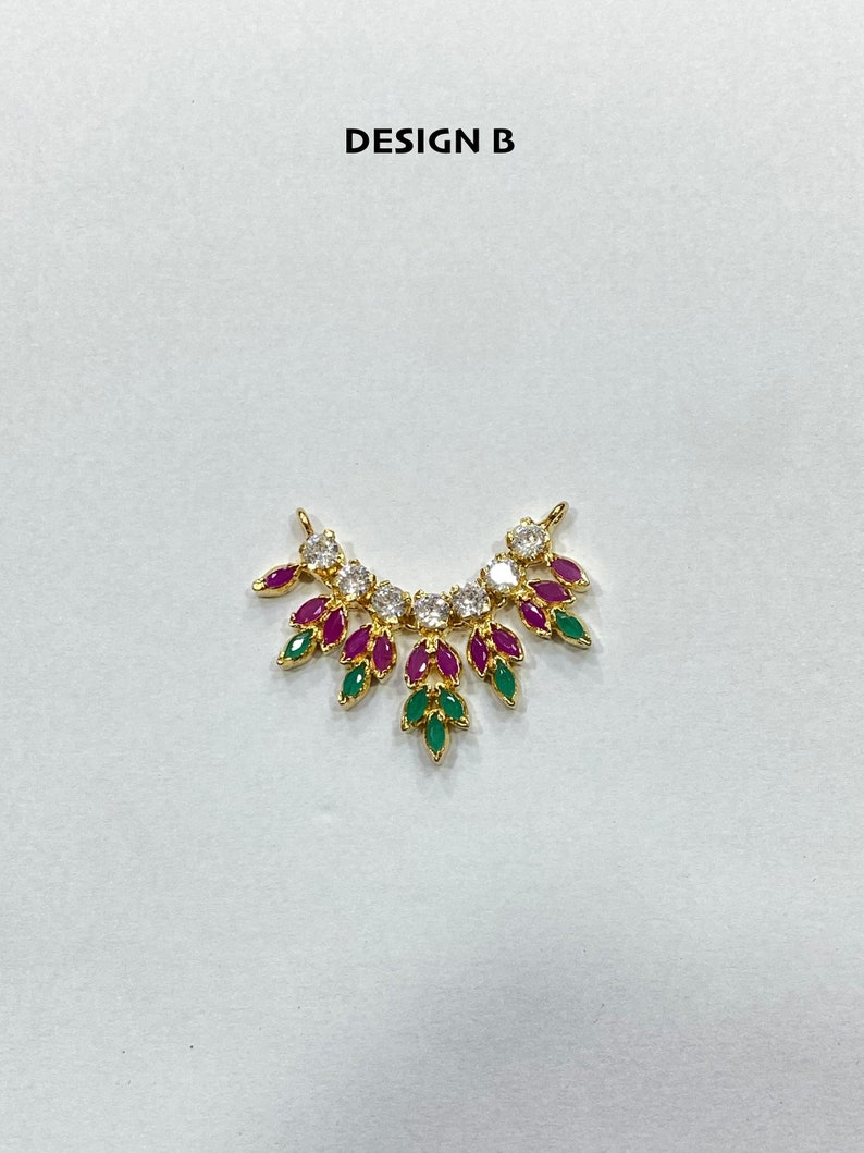 May include: A gold-toned pendant labeled DESIGN B, featuring a curved arrangement of clear round stones and marquise-cut fuchsia and green stones. The piece has two small loops at the top ends for attachment, creating a fan-like, symmetrical jewelry component.