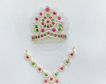 May include: A gold-coloured crown and necklace set. The crown features a floral design with red, green, and clear gemstones. The necklace has a matching design with a central floral pendant. The jewellery is set against a white background.