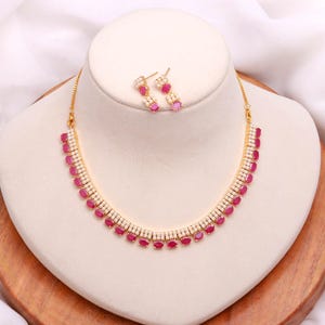 Op de afbeelding: Een gouden ketting- en oorbellenset. De ketting heeft een reeks ovale, robijnkleurige stenen die in een rij zijn geplaatst, afgewisseld met kleinere, witte stenen. De bijpassende oorbellen hebben een vergelijkbaar ontwerp. De sieraden worden op een witte buste getoond.