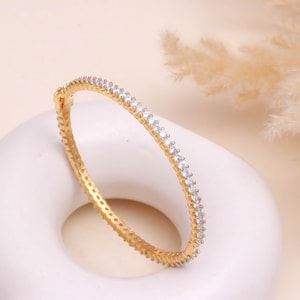 May include: A gold-colored bangle bracelet with a row of small, clear gemstones. The bracelet is set on a white, rounded surface, with a soft, neutral background. The gemstones are evenly spaced along the bracelet's circumference.