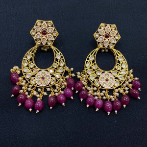 May include: A pair of gold earrings with pink and white enamel floral designs, surrounded by clear stones. The earrings have a crescent moon shape with dangling pink and white beads.