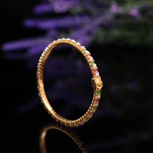 May include: A gold bangle bracelet featuring a row of colourful gemstones and small pearls. The gemstones are in shades of red, green, yellow, and white. The bracelet has a clasp closure and is set against a dark background.