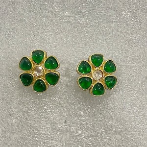 May include: A pair of flower-shaped stud earrings. Each earring features six green, triangular gemstones surrounding a central, clear gemstone. The earrings have a gold-coloured metal setting.