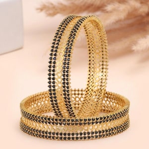 May include: Two gold-toned bangles featuring a central band of intricate openwork heart patterns flanked by two rows of small, round, black stones. The metallic structure has a textured finish, and the design is consistent across both circular bracelets.