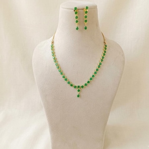 May include: A gold-coloured necklace and earring set. The necklace features a series of green gemstones, with a teardrop pendant. The matching earrings have a similar design, with green gemstones dangling from the ear posts. The jewellery is displayed on a mannequin.