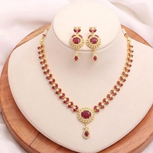 May include: A gold-toned necklace and earring set. The necklace features a large, oval, red gemstone pendant with a smaller teardrop-shaped gemstone below. The earrings and necklace are adorned with smaller red gemstones and white pearls.