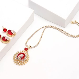 May include: A gold-toned jewelry set consisting of a necklace and matching earrings. Each piece features a large, oval-shaped red gemstone accented with small, clear white stones arranged in a leaf-like pattern and a circular halo setting.
