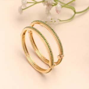 May include: Two gold bangles, each adorned with a line of small green gemstones. The bangles are displayed on a soft, neutral background, with delicate white flowers. The jewellery features intricate detailing.
