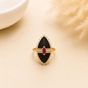 May include: A gold ring featuring a large, marquise-shaped black textured inlay surrounded by a halo of small white stones. A horizontal gold bar crosses the centre, set with a single marquise-cut deep pink stone and small white stones.