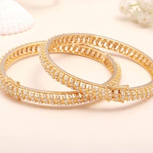 May include: Two gold-coloured bangles with a row of small, white gemstones. The bangles are round and have a clasp closure. The background is a light beige colour, with a seashell and white flowers.
