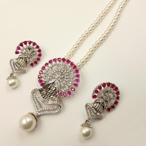 May include: A silver-toned jewelry set featuring a necklace and matching earrings. The necklace has a pearl strand and a pendant with a large white pearl. The pendant and earrings have a floral design with red accents and clear stones.