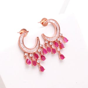 May include: Rose gold-toned crescent earrings with a cascade of pink teardrop-shaped gems. The earrings feature a row of small, clear stones along the crescent curve. A stylish and elegant jewelry piece.