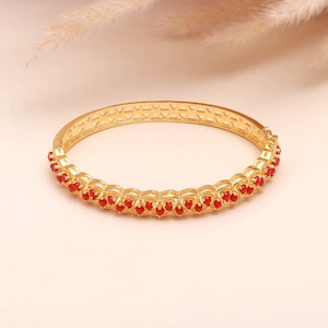 May include: A gold-coloured bangle bracelet featuring a row of small, red gemstones. The bracelet has a geometric design along the top edge. The gemstones are evenly spaced, adding colour to the gold band. This piece of jewellery is a stylish accessory.