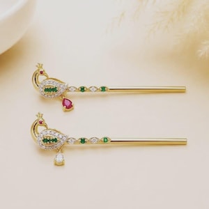 Peut inclure: Deux épingles à cheveux dorées ornées d'un motif de paon. Le corps des paons est incrusté de pierres transparentes et vertes, avec une petite pierre rouge pour l'œil. Une épingle possède une pierre rouge en forme de goutte, l'autre une claire.