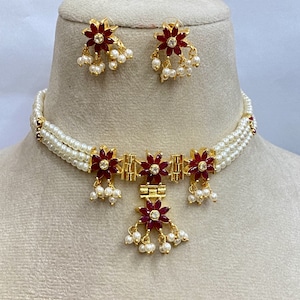 May include: A gold necklace with a red flower pendant and pearl accents. The necklace is paired with matching earrings with red flower accents and pearl drops.