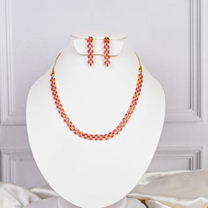 Op de afbeelding: Een gouden ketting- en oorbellenset. De ketting heeft een dubbele rij met afwisselende rode edelstenen. De bijpassende oorbellen zijn bungelend, met een vergelijkbare edelsteenopstelling. De sieraden worden gepresenteerd op een witte buste tegen een zachte achtergrond.