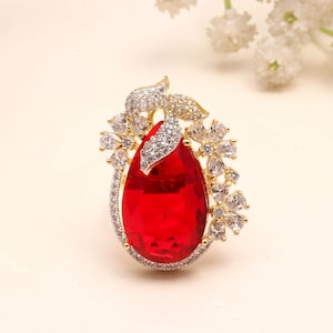 Puede incluir: Un anillo de tono dorado con una gran piedra preciosa roja en forma de pera en el centro. El engaste está adornado con numerosas piedras pequeñas, transparentes y brillantes dispuestas en patrones florales y formas de hojas alrededor de la piedra.