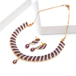 Puede incluir: Un conjunto de collar y pendientes en tono dorado con eslabones curvos y segmentados incrustados con filas alternas de piedras blancas transparentes y magenta intenso. El collar incluye un colgante central en forma de lágrima.