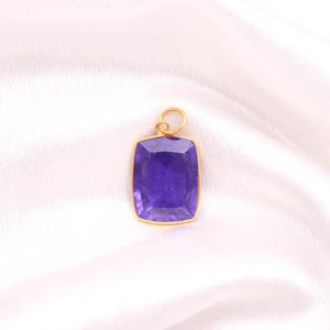 Puede incluir: Un colgante rectangular con una piedra preciosa facetada de color púrpura intenso engastada en un marco de metal fino de tono dorado. El colgante incluye una pequeña anilla circular de tono dorado en la parte superior para sujetarlo a una cadena.