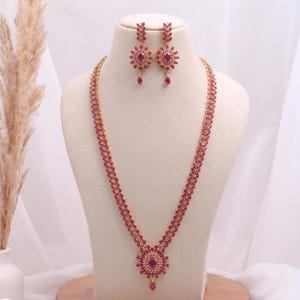 Puede incluir: Un conjunto de collar y pendientes en tono dorado con piedras preciosas de color rubí. El collar presenta una larga cadena con un colgante central y un par de pendientes a juego. Las joyas se exhiben en un maniquí sobre un fondo blanco.