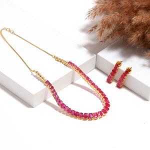 Puede incluir: Un conjunto de collar y pendientes dorados. El collar y los pendientes presentan una serie de piedras preciosas rosas rectangulares y vibrantes. Las joyas se exhiben sobre una superficie blanca, con hierba ornamental seca en el fondo.