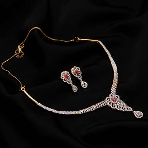 Puede incluir: Un conjunto de collar y pendientes en tono dorado con piedras transparentes en pavé y piedras centrales rojas en forma de lágrima. El collar presenta un diseño de colgante central con un elemento colgante, a juego con los pendientes.